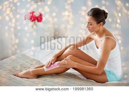 beauty, depilation, epilation, hair removal and people concept - beautiful woman applying depilatory wax strip to her leg skin at home bedroom