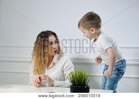Young Woman Secretary With Her Little Son In The Workplace In Office. Child Requires His Mother To P