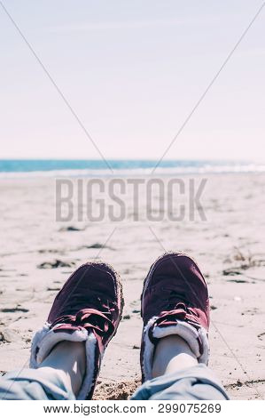 Feet Of Young Woman In Sport Footwear And Jeans Relaxing After Hiking On Sand Beach. Pov. Adventure 
