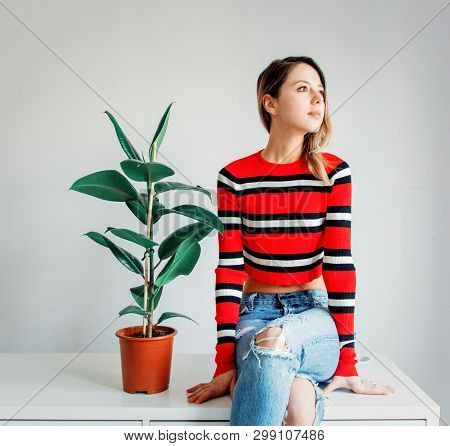 Young Woman In Sptriped Sweater And Jeans Sitting On A Table Near Ficus