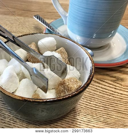 Bowl With Pieces Of White And Brown Sugar Cubes And With Sugar-tongs On A Wooden Table In Front Of A