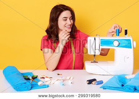 Indoor Shot Of Charming Smiling Woman Putting Thread On Sewing Machine, Being In Good Mood, Likes He