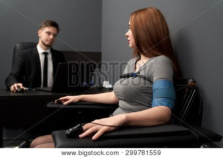 Woman Sitting In Chair With Attached Finger Sensors And Indicators, Looking At Lie Detector. Young M