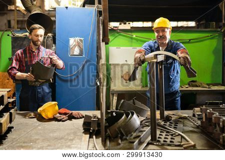 One of two contemporary machinebuilders preparing metallic workpieces for processing while his colleague going to put on welding mask