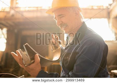 Aged successful engineer or technician in hardhat, gloves and uniform looking at metallic semi-circle detail in his hands
