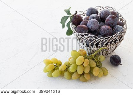 Just Picked Ripe Plums In Wicker Basket And A Bunch Of Ripe White Grapes On The White Structured Bac