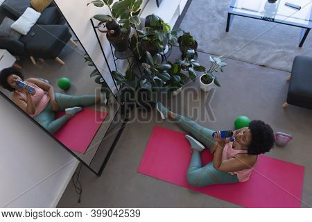 Overhead view of african american woman sitting on exercise mat drinling water. self isolation fitness at home during coronavirus covid 19 pandemic.