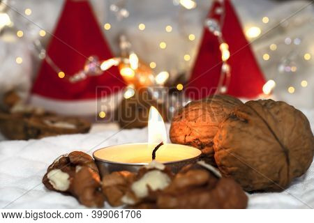 A Burning Candle Stands Near Walnuts Against A Background Of Blurry Lights And Two Santa Claus Hats