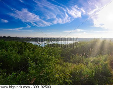 Pond Seen From Hill Surrounded By Wooded Forest And Green Foliage And Fauna With Bright Blue Sky And