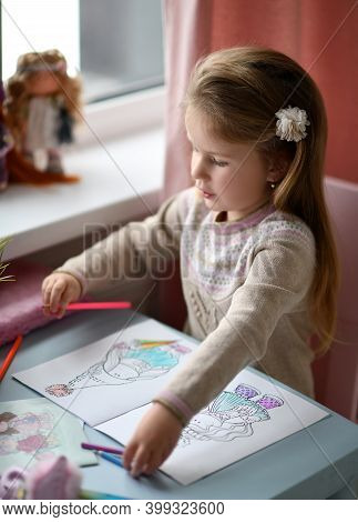Pretty Small Concentraited Girl Child In Sweater Sitting And Coloring Doll Drawing In Album With Pen