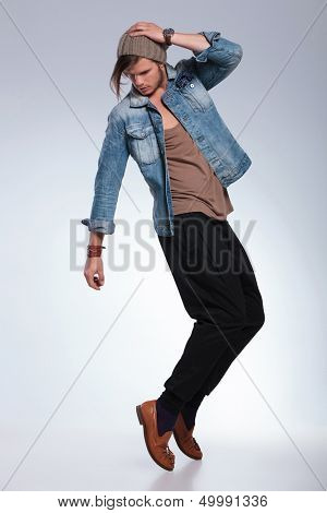full length portrait of a casual young man balancing on his toes while looking down and holding his hand on his head. on gray studio background