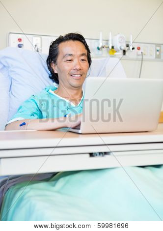 Happy mature male patient using laptop on bed in hospital