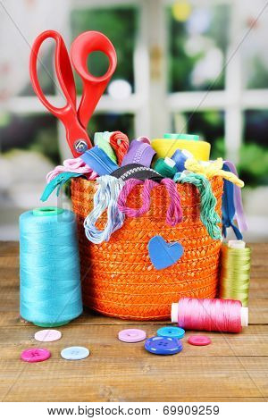 Round orange box full of multicoloured thread and everything necessary for sewing on a brown wooden table on window background
