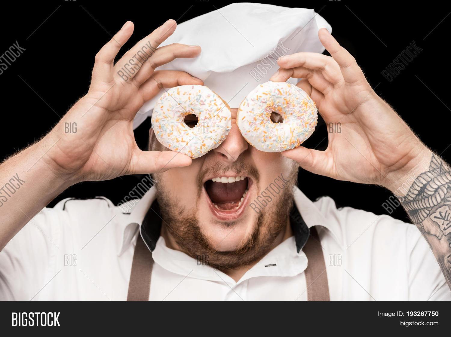 excited chef in hat and apron looking through doughnuts isolated
